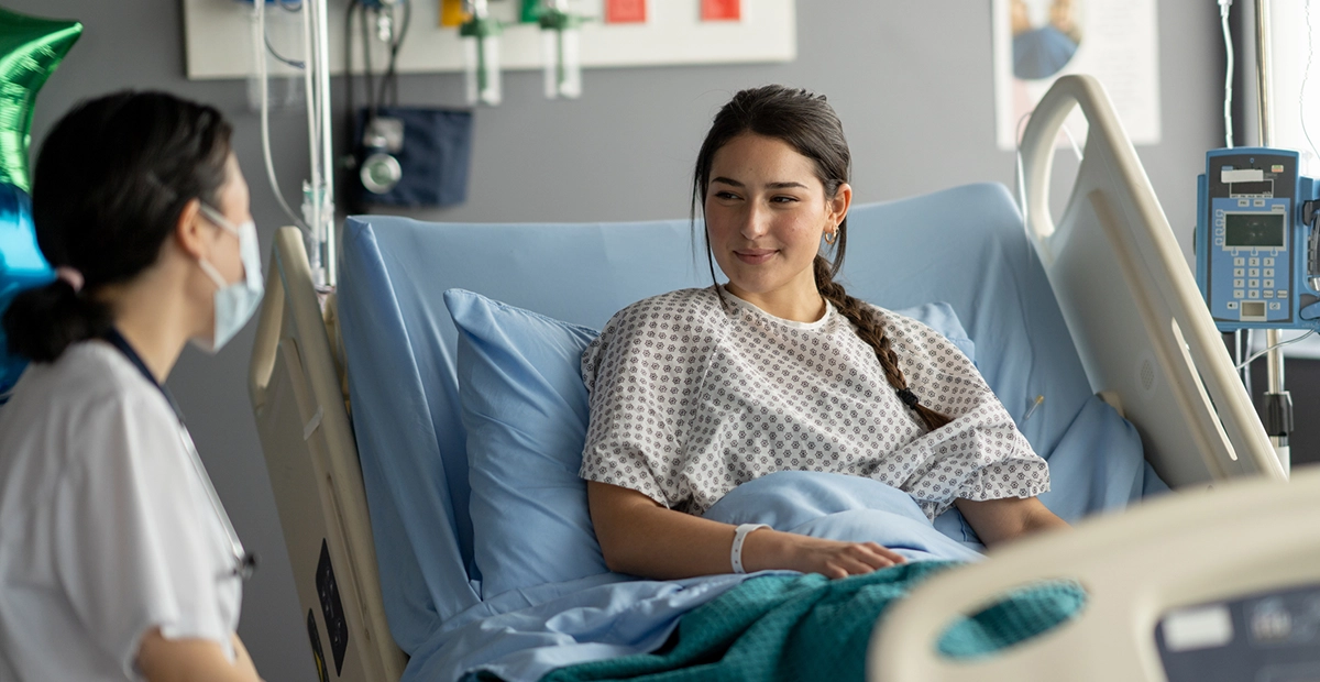 A young female sits up in her hospital bed as she recovers after surgery. She is dressed comfortably in a gown and tucked under warm blankets as she talks with her doctor who is making rounds.