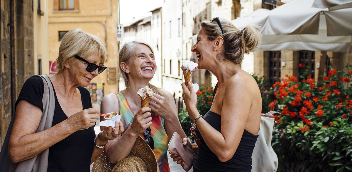 Three mature female friends standing eating Italian ice-creams while in a street in Tuscany during summer