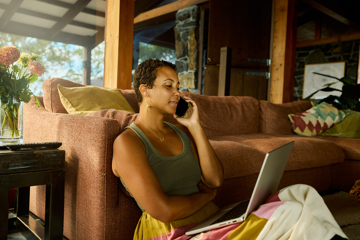 Woman sitting on the floor leaning against her sofa, on the phone with her laptop in her lap